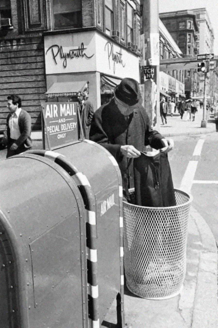 A man in a hat and coat stands on a city sidewalk, reading mail beside an airmail mailbox and a trash can. Pedestrians and buildings are visible in the background.