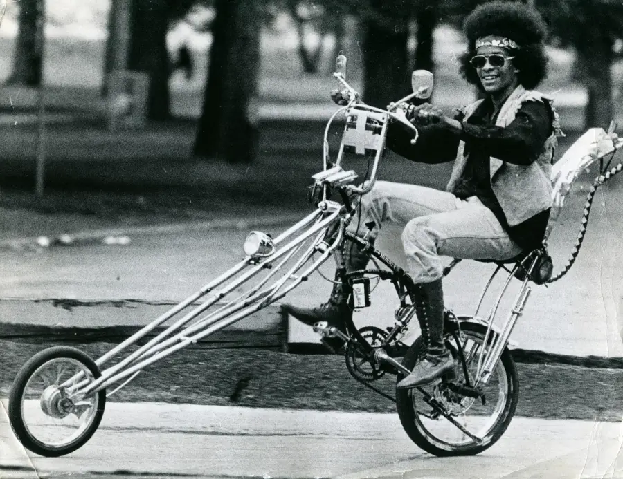 A person with sunglasses and an afro rides a customized tall chopper bicycle with long handlebars and front fork, smiling and raising one hand, in a park setting.