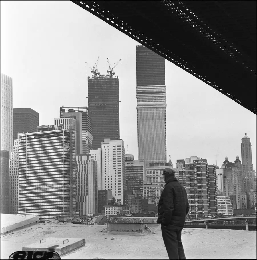 A person stands on a rooftop, looking at New York City’s skyline, with the Twin Towers under construction in the background and a bridge structure overhead.