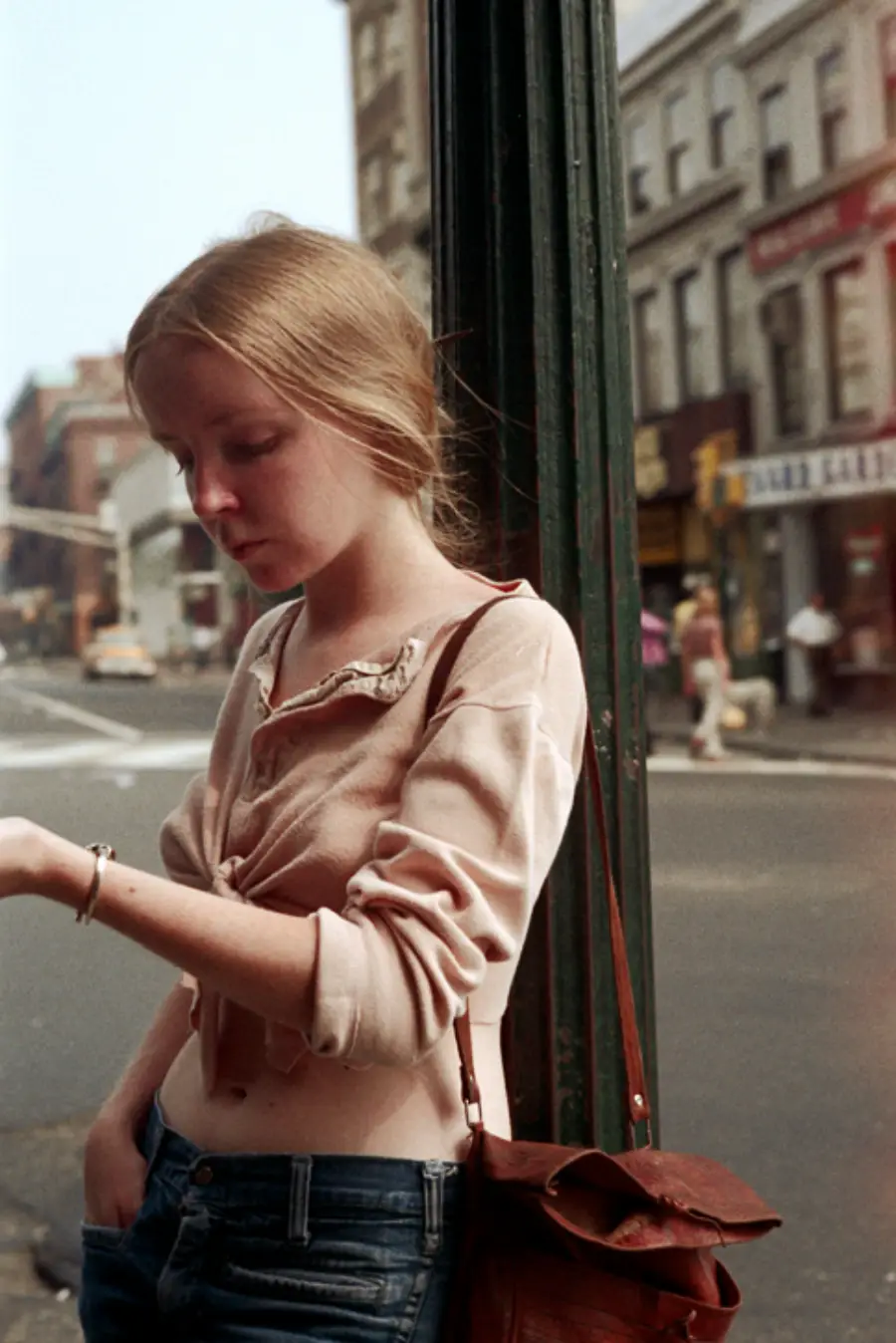A young woman with long blonde hair stands by a pole on a city street, looking down at her hand. She wears a light pink, tied-up shirt and jeans. Buildings, people, and shops are visible in the background.