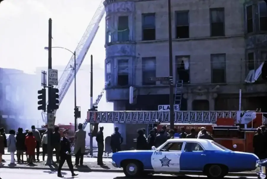A crowd watches firefighters respond to a building fire with a ladder truck. Smoke is visible near the building, and a blue police car is parked at the scene.