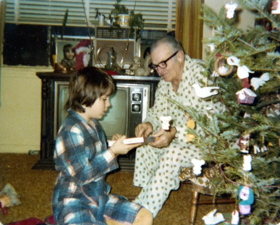 A child and an elderly man in pajamas sit by a Christmas tree, exchanging a small gift in a cozy, vintage living room with a TV and family photos in the background.