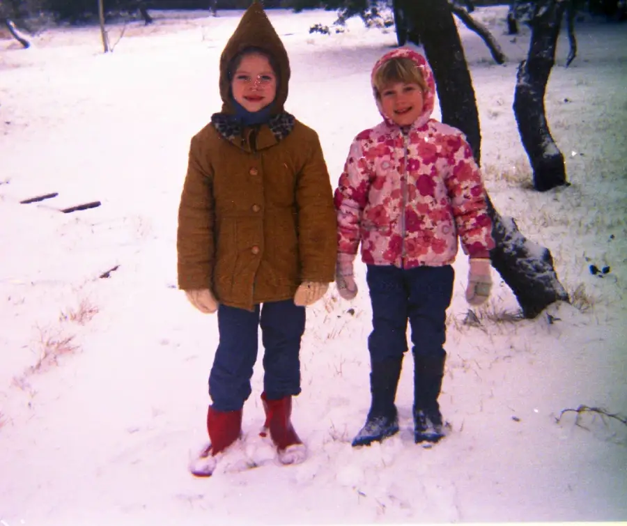 Two children wearing winter coats, boots, and gloves stand together on snow-covered ground with trees in the background, smiling at the camera.