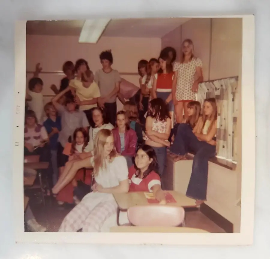 A group of teens pose playfully in a classroom, some sitting on desks and others standing or sitting on a countertop by the window. Everyone appears relaxed and casual, with various expressions and outfits.