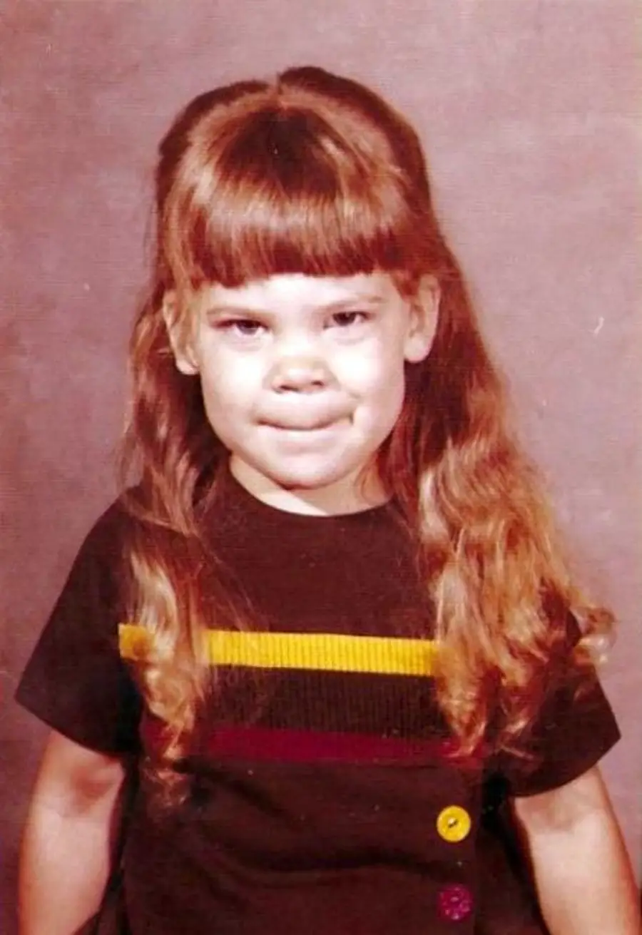 A young girl with long, wavy hair and bangs wears a brown dress with red, yellow, and orange stripes and buttons. She smiles slightly and stands in front of a plain, brownish background.