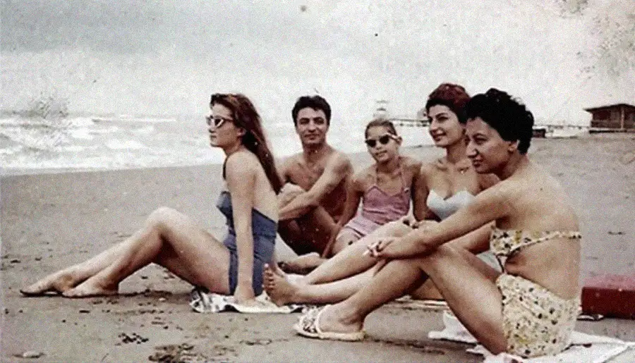 Five people in swimsuits sit and relax on a sandy beach with waves in the background under a cloudy sky. The group includes three women in the foreground, one woman and one man behind them.