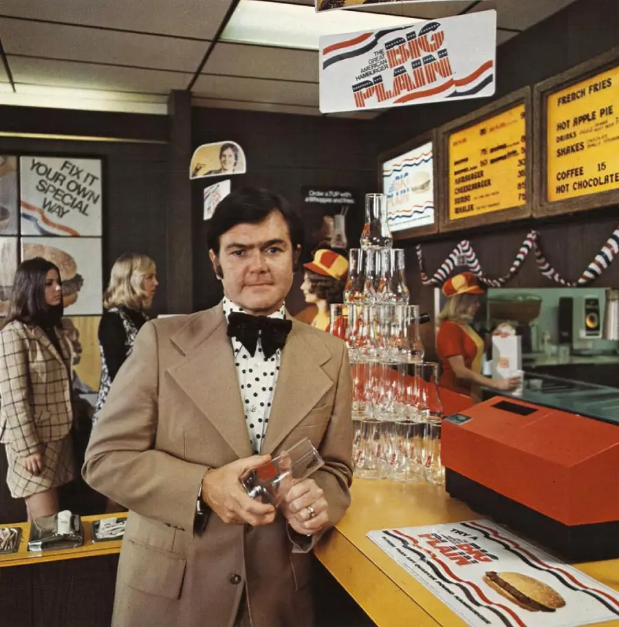 A man in a tan suit with a polka dot bow tie stands by a counter in a retro fast food restaurant, holding a glass. Behind him are stacked glasses, women in uniforms, and menu boards with red, white, and blue décor.