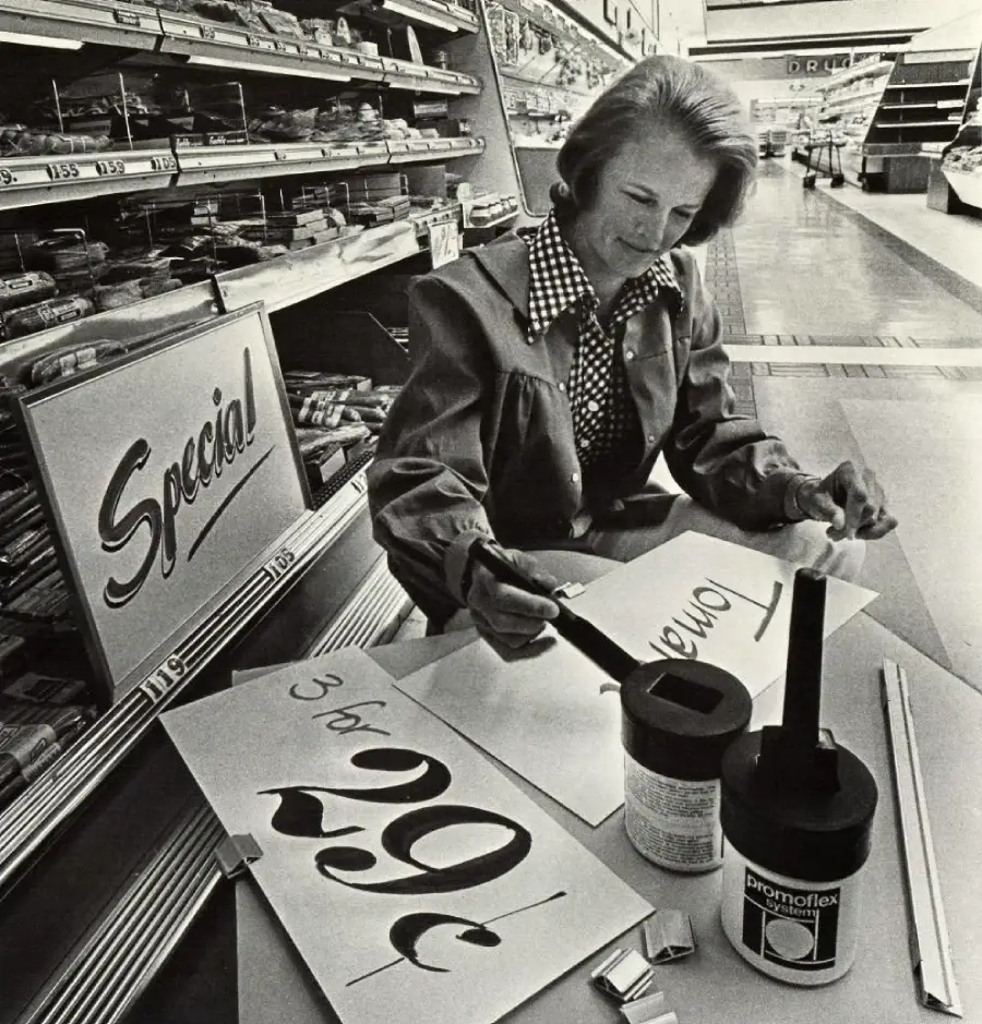A woman sits at a table in a grocery store aisle, hand-lettering sale signs with black ink. One sign reads “3 for 29¢.” Shelves of products line the background.