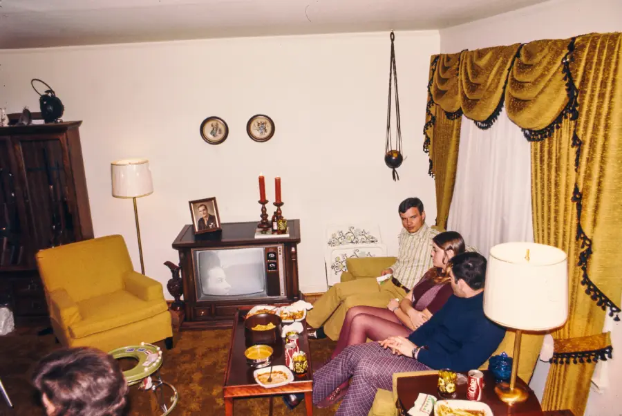 Four adults sit and talk in a vintage living room with gold curtains, yellow armchairs, a box TV, and snacks on tables. A photo and candles sit on the TV. The scene appears to be from the 1970s.