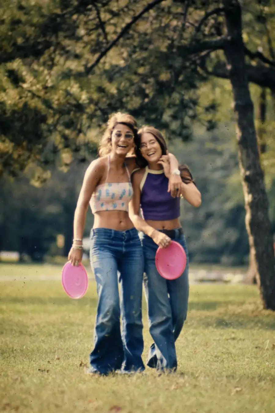 Two smiling women holding pink frisbees walk arm in arm on green grass in a park, surrounded by trees, both wearing jeans and crop tops.
