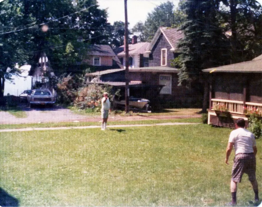 Two people play catch on a grassy lawn in front of houses and trees. A car is parked in the driveway in the background under a partly sunny sky.