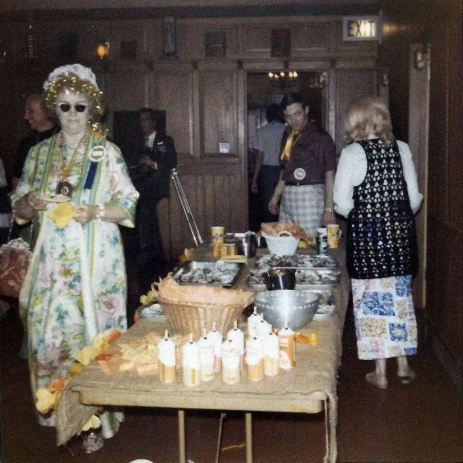 People stand around a table filled with food and drink in a wood-paneled room. A person in colorful, floral attire with sunglasses and a hat stands on the left, while others are near the table.