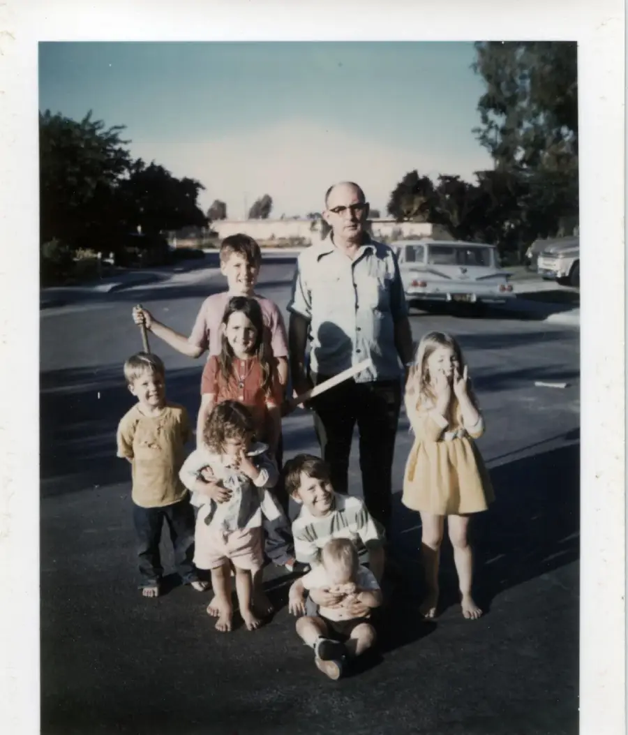 A man stands on a street surrounded by seven children. Some children are sitting, some are standing, and all are barefoot. There are cars and trees in the background under a clear sky.