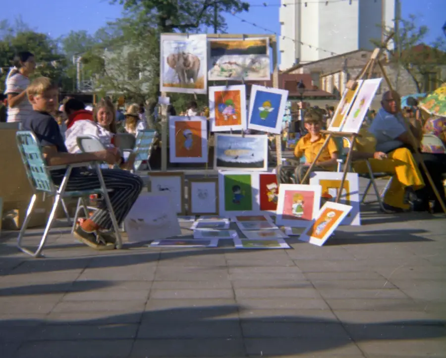 People sit on folding chairs at an outdoor art fair. Various colorful paintings are displayed on easels and on the ground. Other people and buildings are visible in the background. The scene is sunny and lively.