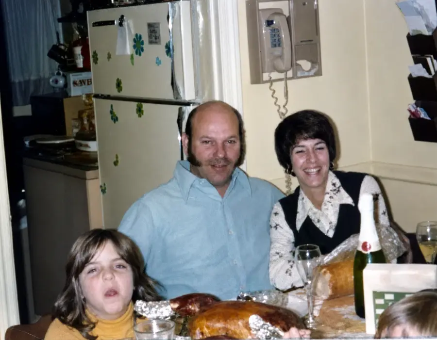 Three people sit at a kitchen table with food, including roast meats and a bottle, smiling for the camera. A refrigerator with magnets and an old wall phone are visible in the background.