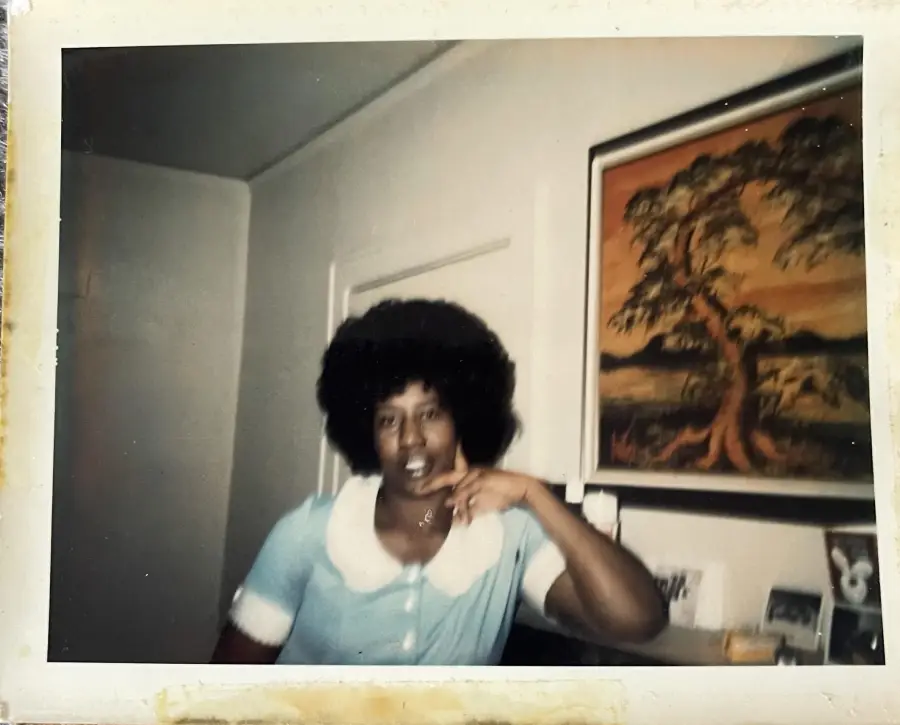 A person with an afro hairstyle in a blue dress with a white collar poses indoors near a wall with a framed picture of a tree.