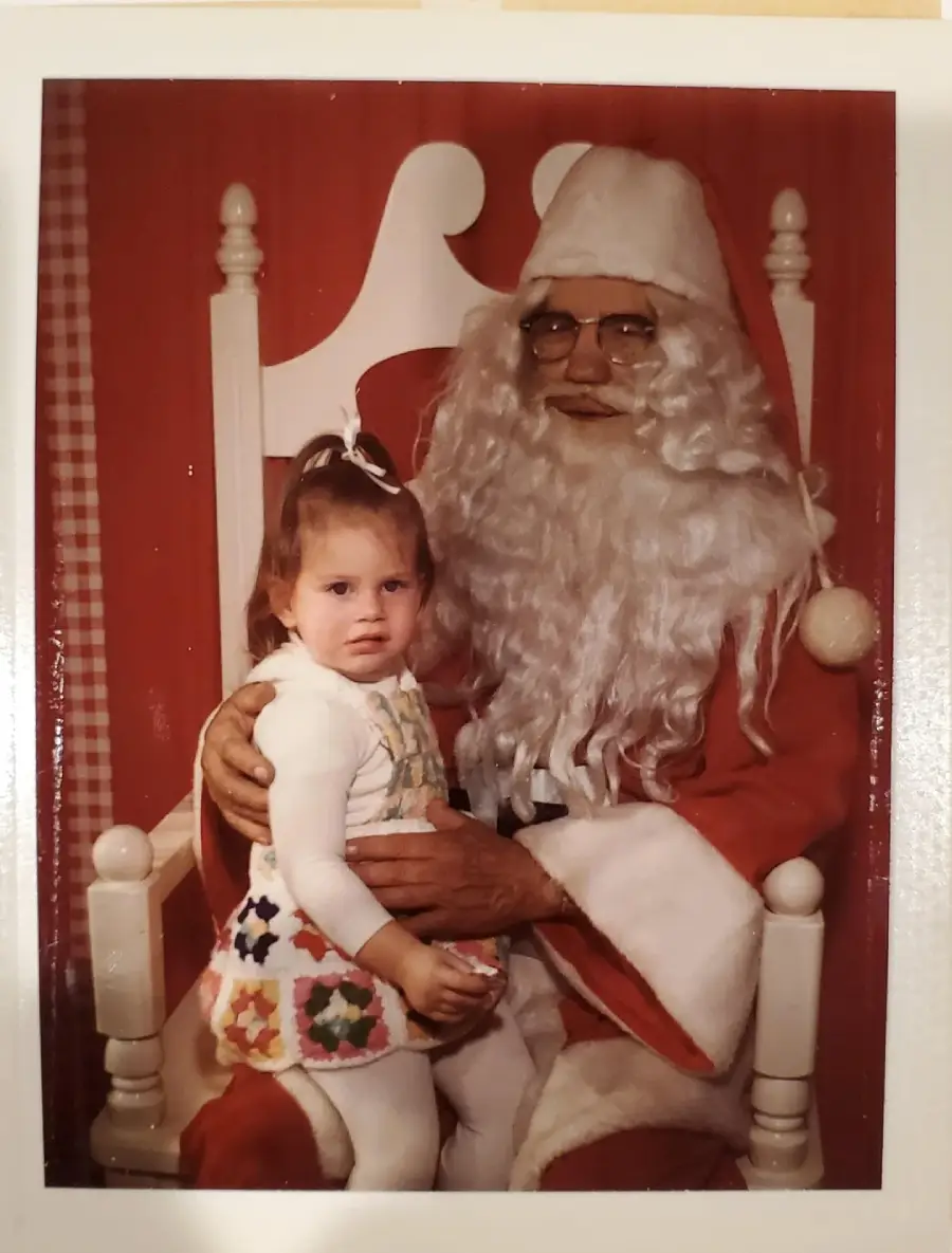 A young child sits on Santa’s lap in a festive setting with red and white décor. Santa wears glasses, a red suit, and a white beard. The child looks slightly uneasy but is dressed neatly with a bow in her hair.