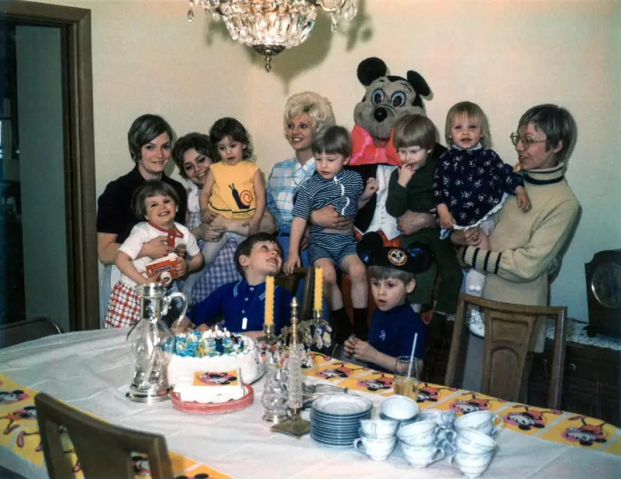 A group of children and adults gather around a table with a birthday cake. Some kids wear party hats; one person is in a cartoon mouse costume. The room is decorated, and everyone appears to be celebrating.