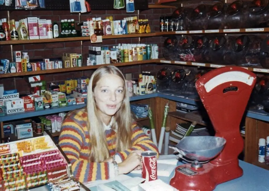 A woman with long blonde hair sits behind a pharmacy counter with shelves of products, a red scale, and a Coca-Cola can in front of her.