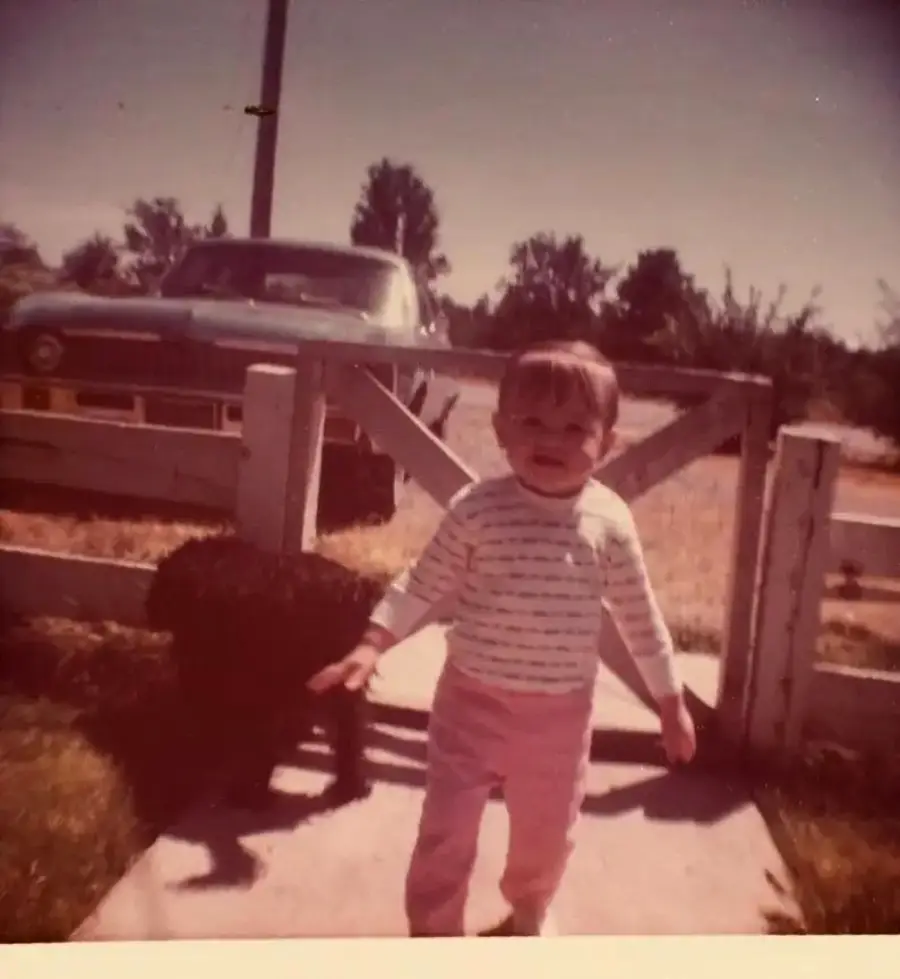 A young child in striped shirt and pink pants stands on a walkway near a wooden gate, with a vintage car parked in the background on a sunny day.