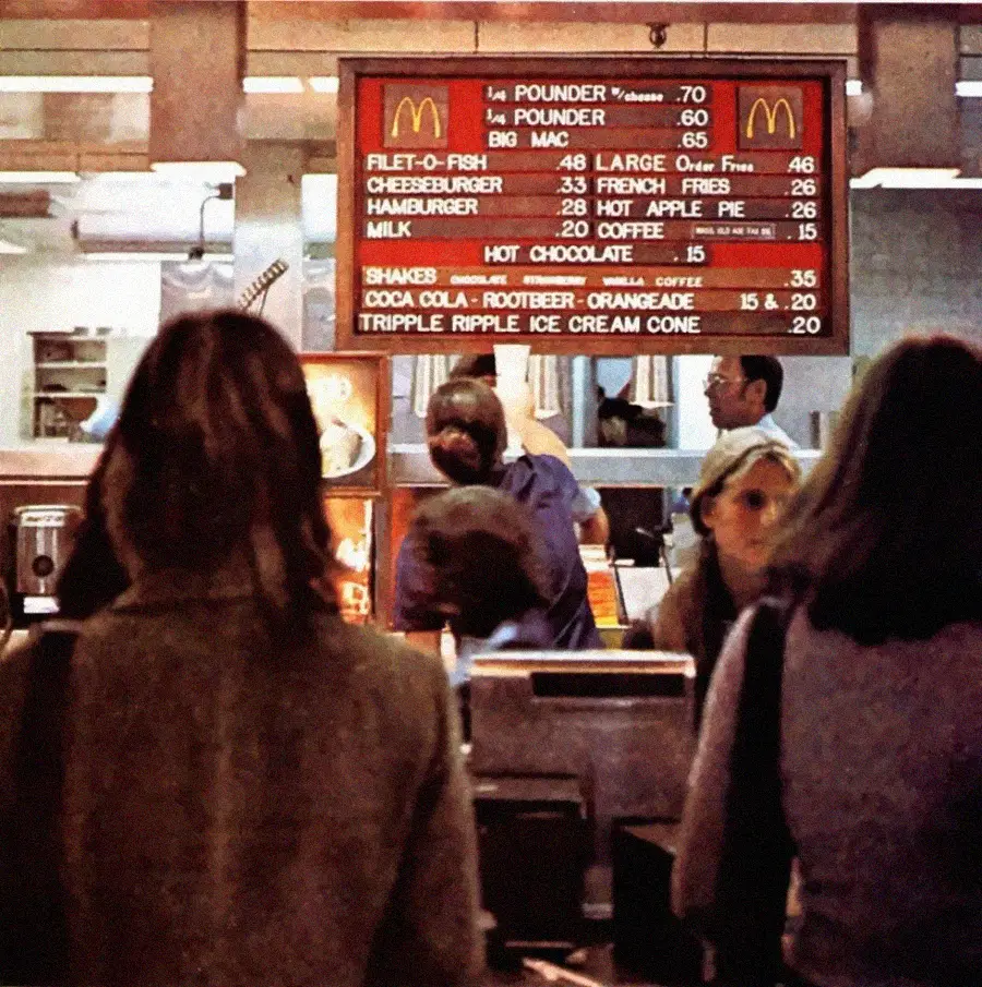 People stand at a McDonald’s counter with a retro menu board overhead displaying classic items and prices. The scene has a vintage atmosphere, likely from the 1970s.