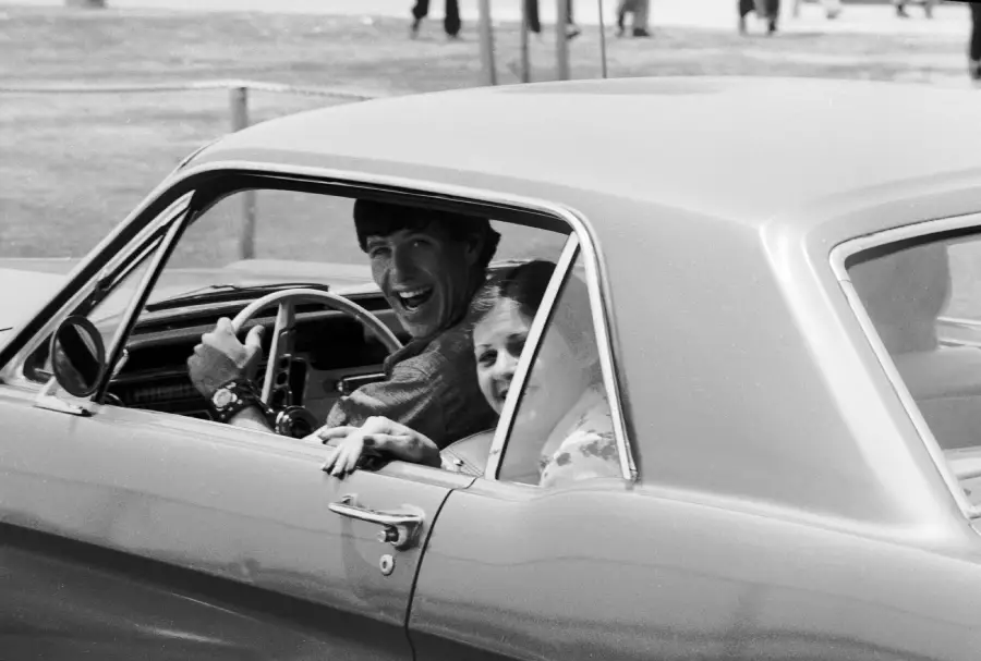 A man and a young girl smile while sitting in the front seats of a classic car with the window down. The man is in the driver’s seat and both look toward the camera. The background shows grass and people walking.