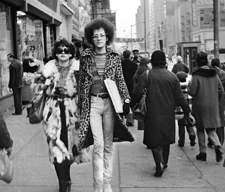 Two people in patterned coats and sunglasses walk arm-in-arm down a busy city sidewalk in the 1970s, surrounded by pedestrians and storefronts.