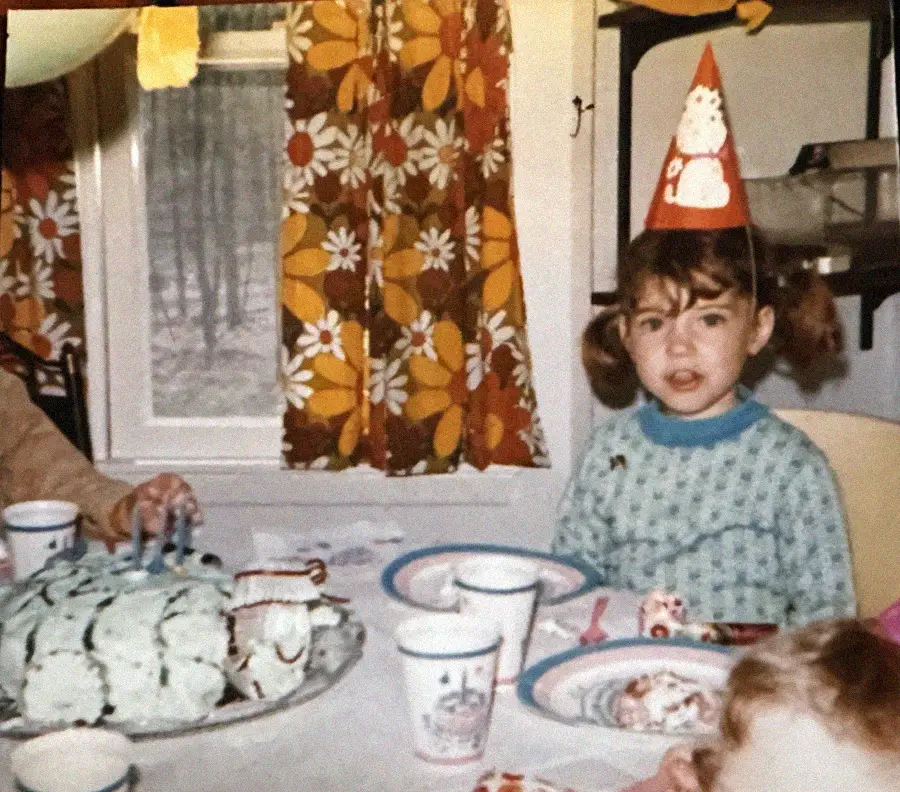 A young girl in a party hat sits at a table with a blue cake, paper plates, and cups. Colorful, floral curtains and a window are in the background. Another child and an adult’s hand are partially visible.