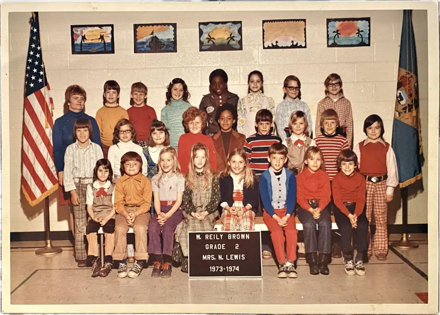 A class photo of 24 children and one adult, with an American flag and class sign reading “W. Reily Brown Grade 2, Mrs. N. Lewis, 1973-1974.” Children’s artwork is displayed on the wall behind them.