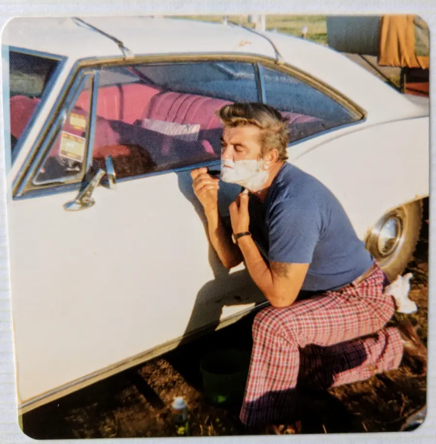 A man in plaid pants kneels by a white car, shaving his face with shaving cream, using the car’s window as a mirror. The car has a pink interior and is parked outdoors.