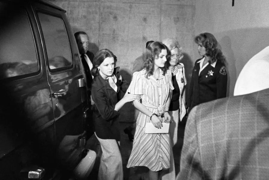 A young woman in a striped dress is escorted by officers and surrounded by people as she walks near a dark van in a dimly lit area.