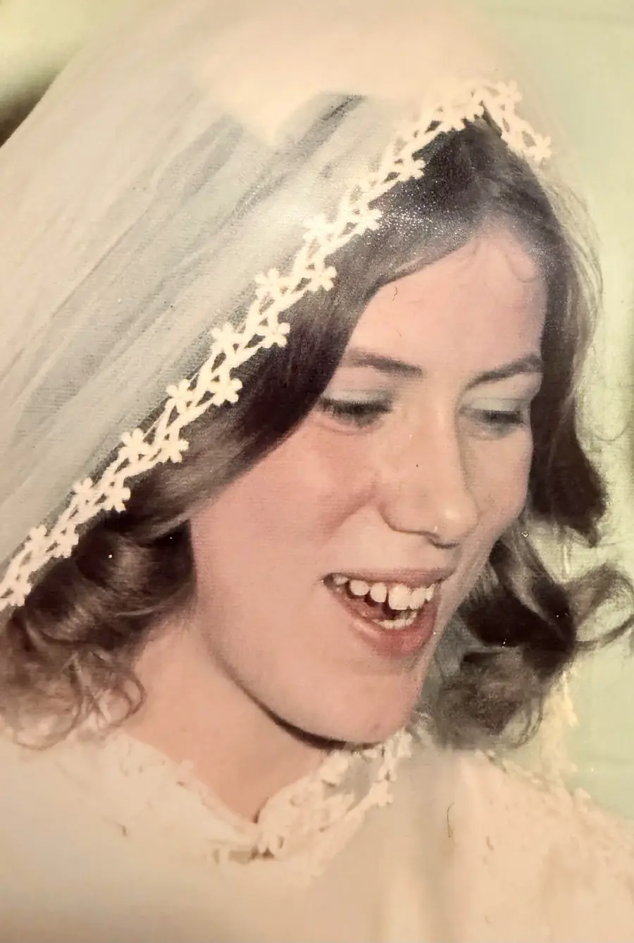 A young woman in a white wedding veil with lace trim smiles while looking down. She has wavy brown hair and is wearing a white dress. The background is out of focus.