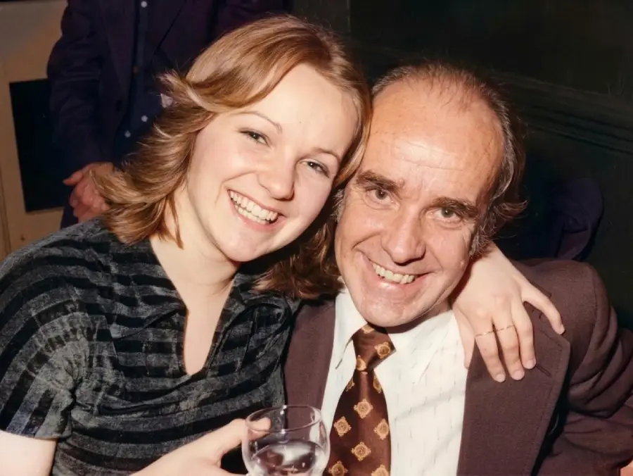 A young woman with blonde hair smiles and hugs an older man in a suit and patterned tie. Both are seated and smiling at the camera; the man holds a glass of water.