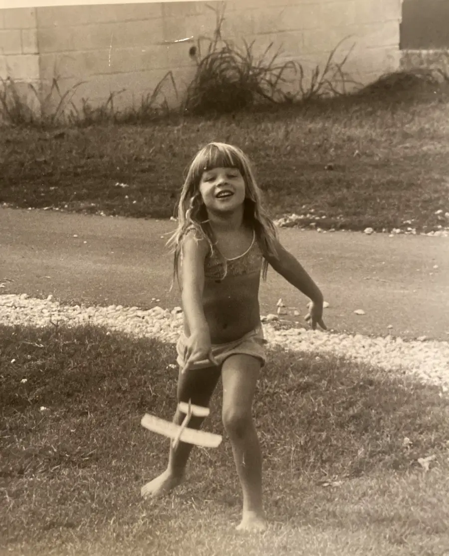 A young girl runs barefoot on grass, smiling and holding a small toy airplane. The photo is black and white, with a building and pathway in the background.