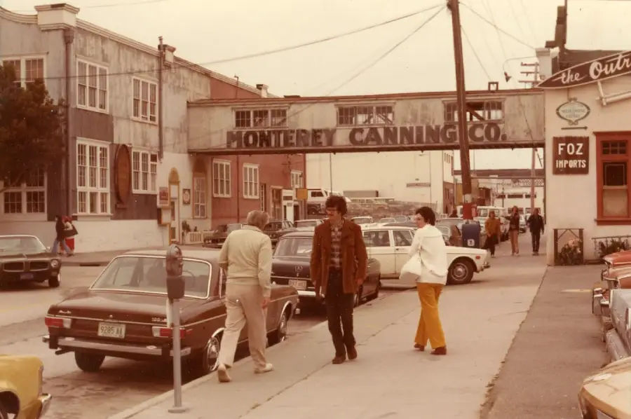Three people walk near parked cars on a street below a sign reading “Monterey Canning Co.” The scene appears to be from the 1970s, with vintage cars and buildings lining both sides of the street.