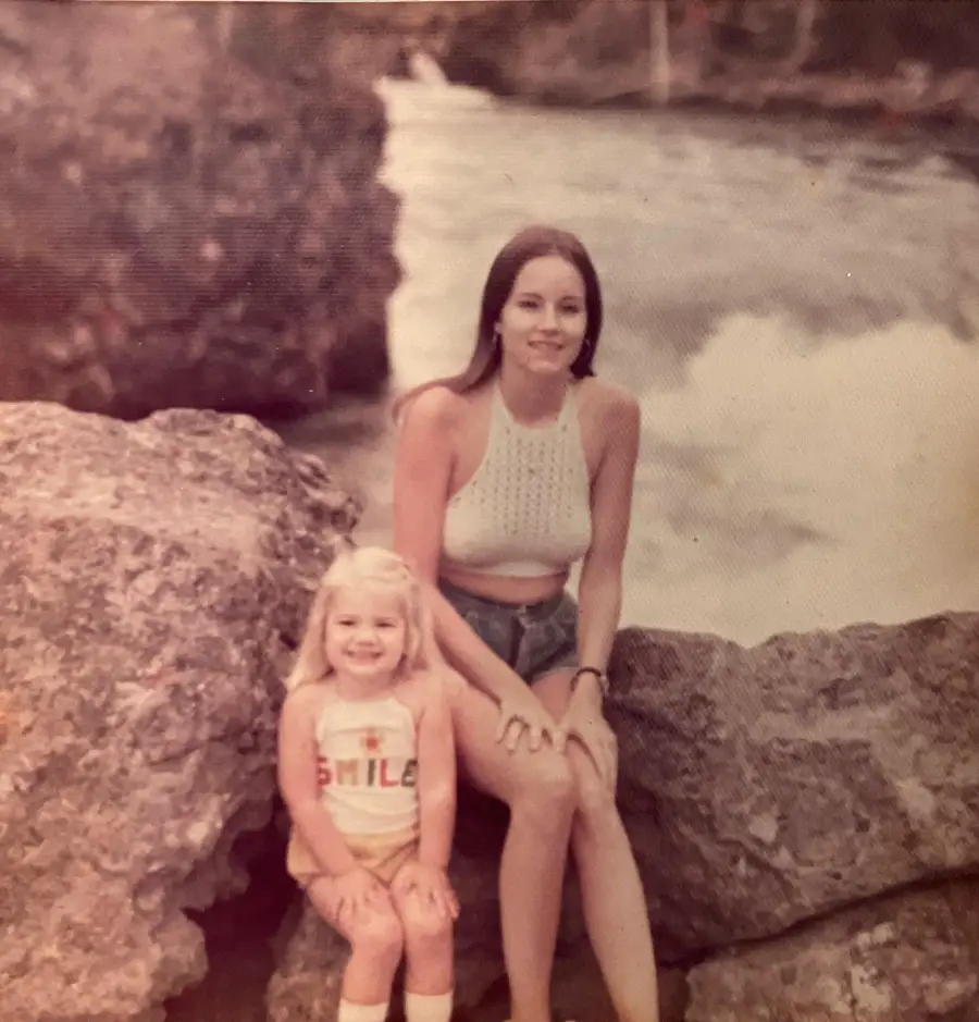A woman and a young girl sit on large rocks by a river. The woman wears a white crocheted top and shorts, and the girl wears a shirt with the word “SMILE” and smiles at the camera. Trees and water are in the background.