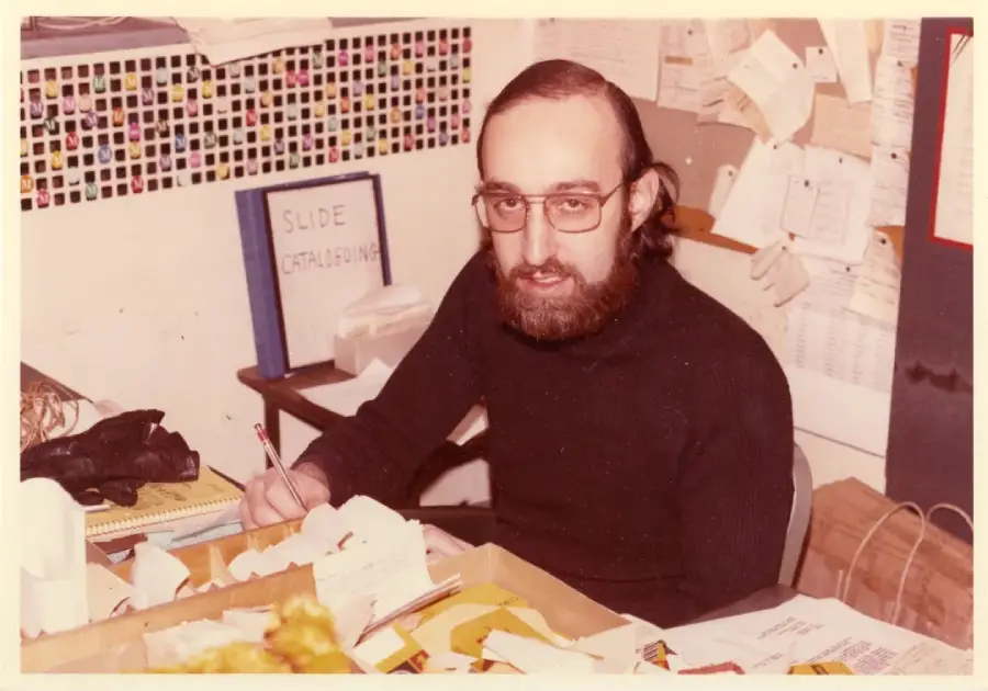 A bearded man with glasses sits at a cluttered desk, writing on paper. Behind him is a wall covered with notes and a binder labeled “Slide Cataloging.” The setting appears to be an office or workspace.