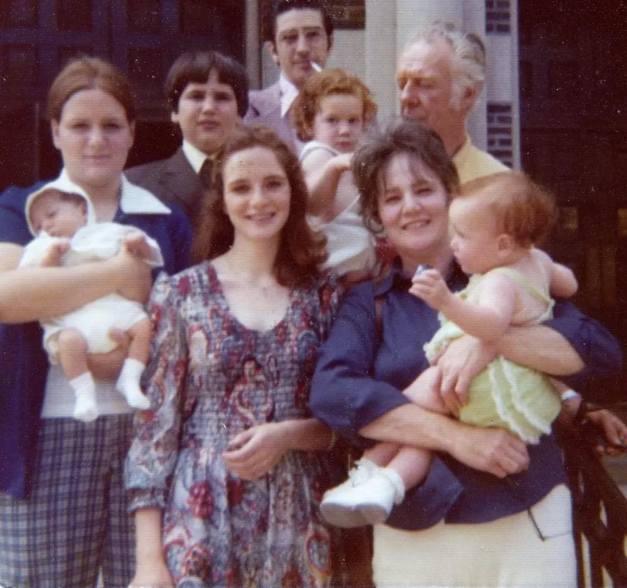 A vintage photo of seven people posing outdoors: three women, two men, and two babies. One woman holds a baby in white, another holds a baby in yellow. They all appear to be smiling or neutral, standing in front of a building.