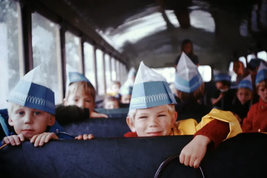 Children wearing blue and white paper hats sit on a school bus, smiling and looking toward the camera. The bus is filled with kids, and the atmosphere appears cheerful and lively.