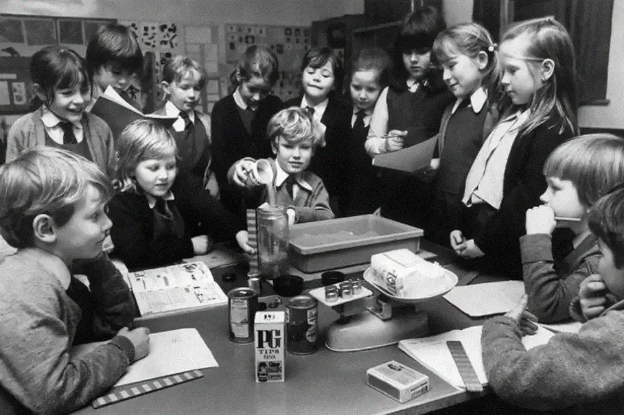A group of children in school uniforms gathers around a table, watching as one child stacks canned goods. A scale, tea box, and other food items are on the table. The classroom has drawings on the walls.