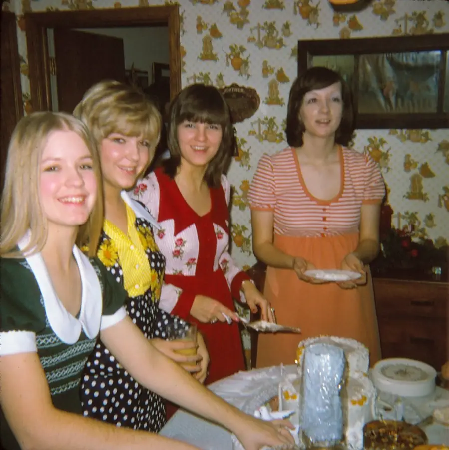 Four women smile at a table set with food and cake, in a vintage kitchen with patterned wallpaper. One woman is serving cake, and others stand close together, appearing to enjoy a gathering or celebration.