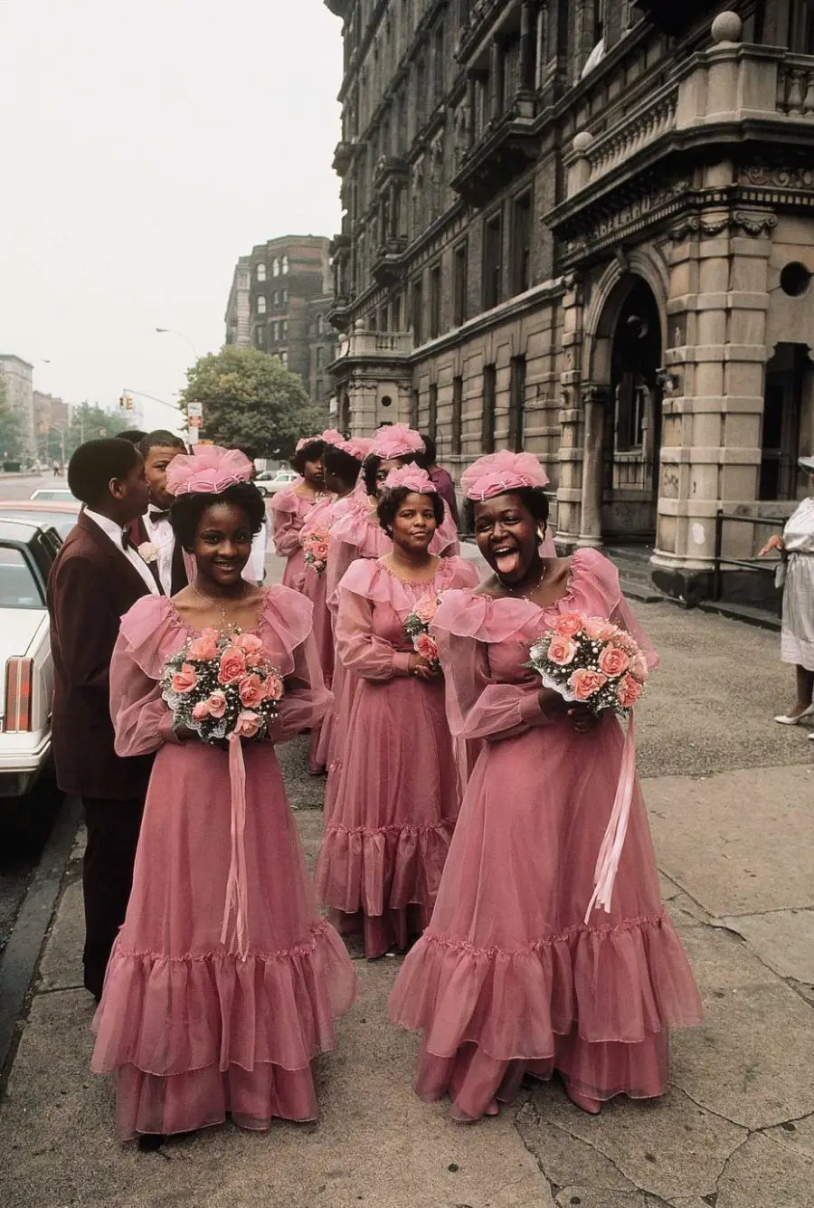 A group of bridesmaids in matching pink dresses and floral headbands stand outside a city building, holding bouquets and smiling, with other wedding guests nearby.