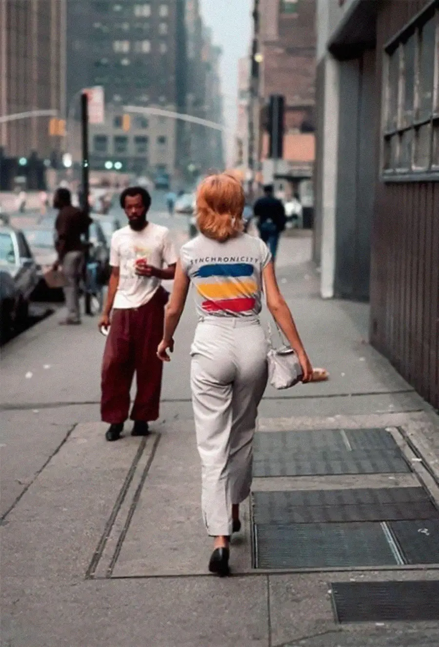 A woman with red hair walks down a city sidewalk, wearing white pants and a colorful “SYNCHRONICITY” T-shirt. A man stands nearby, holding a drink. Tall buildings and cars line the busy urban street.