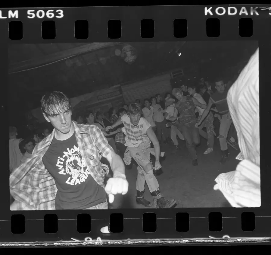 Black-and-white photo of people dancing energetically at a concert. The person in front wears a plaid shirt over a t-shirt with “ANTI NOWHERE LEAGUE” text. The crowd behind appears animated and engaged.
