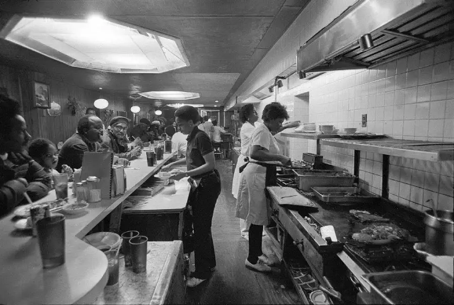 Black and white photo of a busy diner kitchen. Cooks work at the stove while customers sit at the counter, some eating and others waiting. The scene is lively, with visible grills, plates, and drinks.