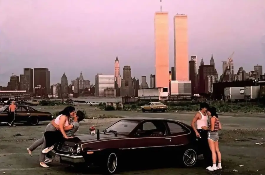 Three people stand by a car in a lot at sunset, with two kissing on the hood. The New York City skyline and Twin Towers are visible in the background.