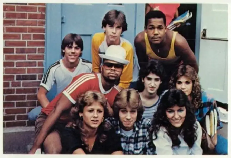 Nine teenagers pose together outside a building, some smiling, dressed in casual 1970s or 1980s clothing. The group includes both young men and women with various hairstyles, and a brick wall is visible in the background.
