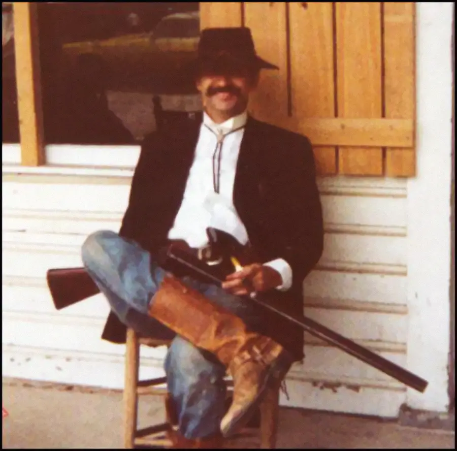 Man in western attire with a hat, boots, and mustache, sits on a chair holding a rifle. He’s wearing a black coat, white shirt, and bolo tie. A wooden shutter and window are behind him.