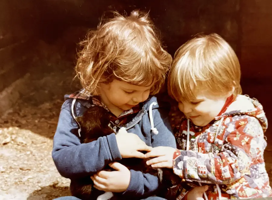 Two young children sit closely together, smiling and gently holding a small black and white puppy in their arms on a sunny day.