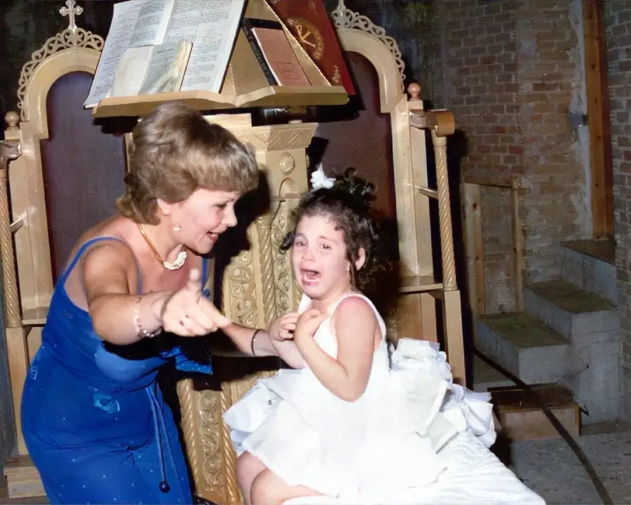 A woman in a blue dress gestures and smiles while a young girl in a white dress sits and cries in front of an ornate wooden lectern with an open book.